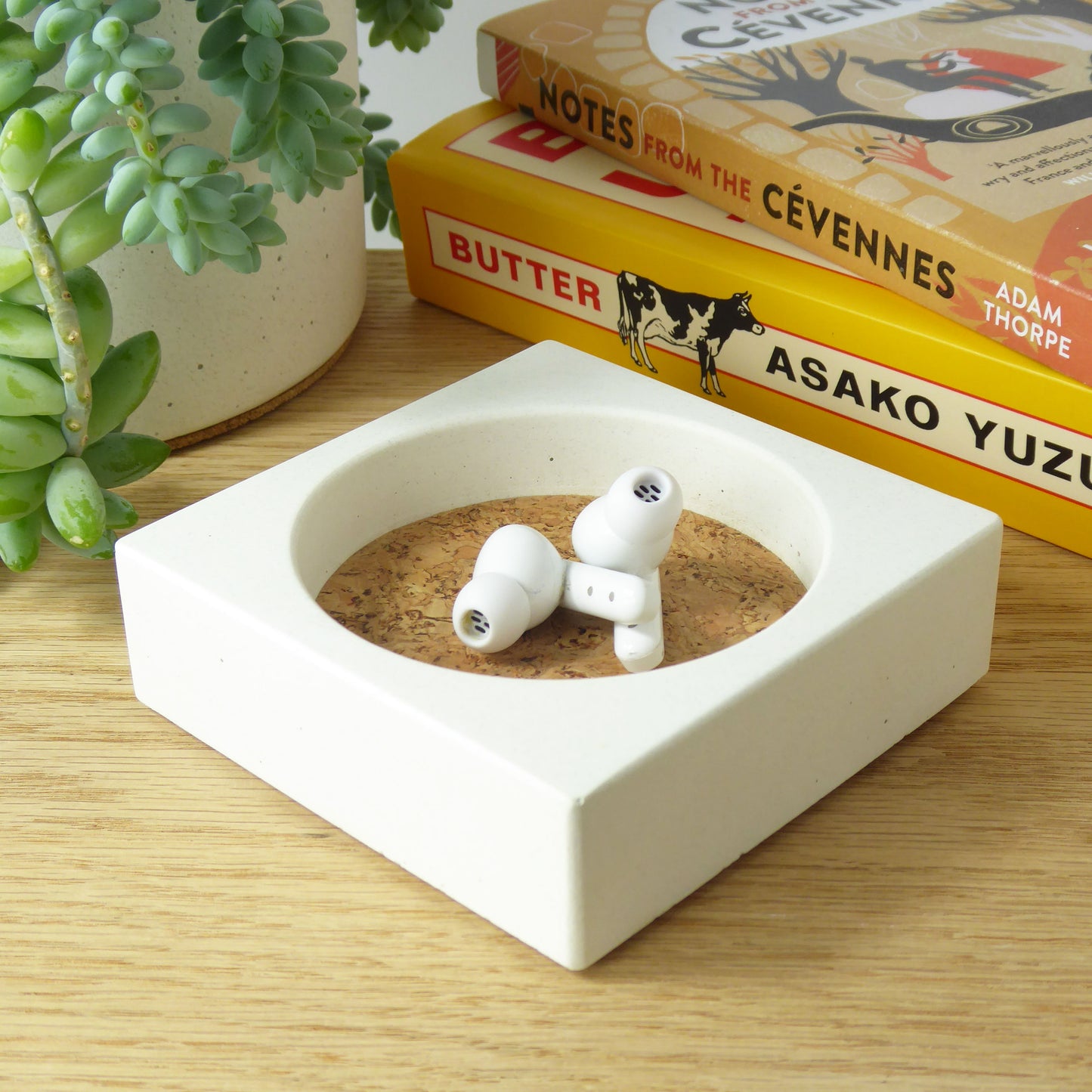 White concrete trinket tray with ear buds inside on a wooden shelf with books and a plant in the background.
