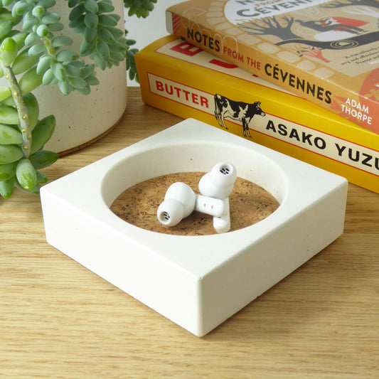 White concrete trinket tray with ear buds inside on a wooden shelf with books and a plant in the background.