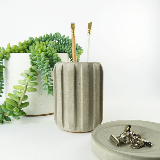 Grey concrete textured pen holder with pencils in it, a small plant and a tray with paperclips