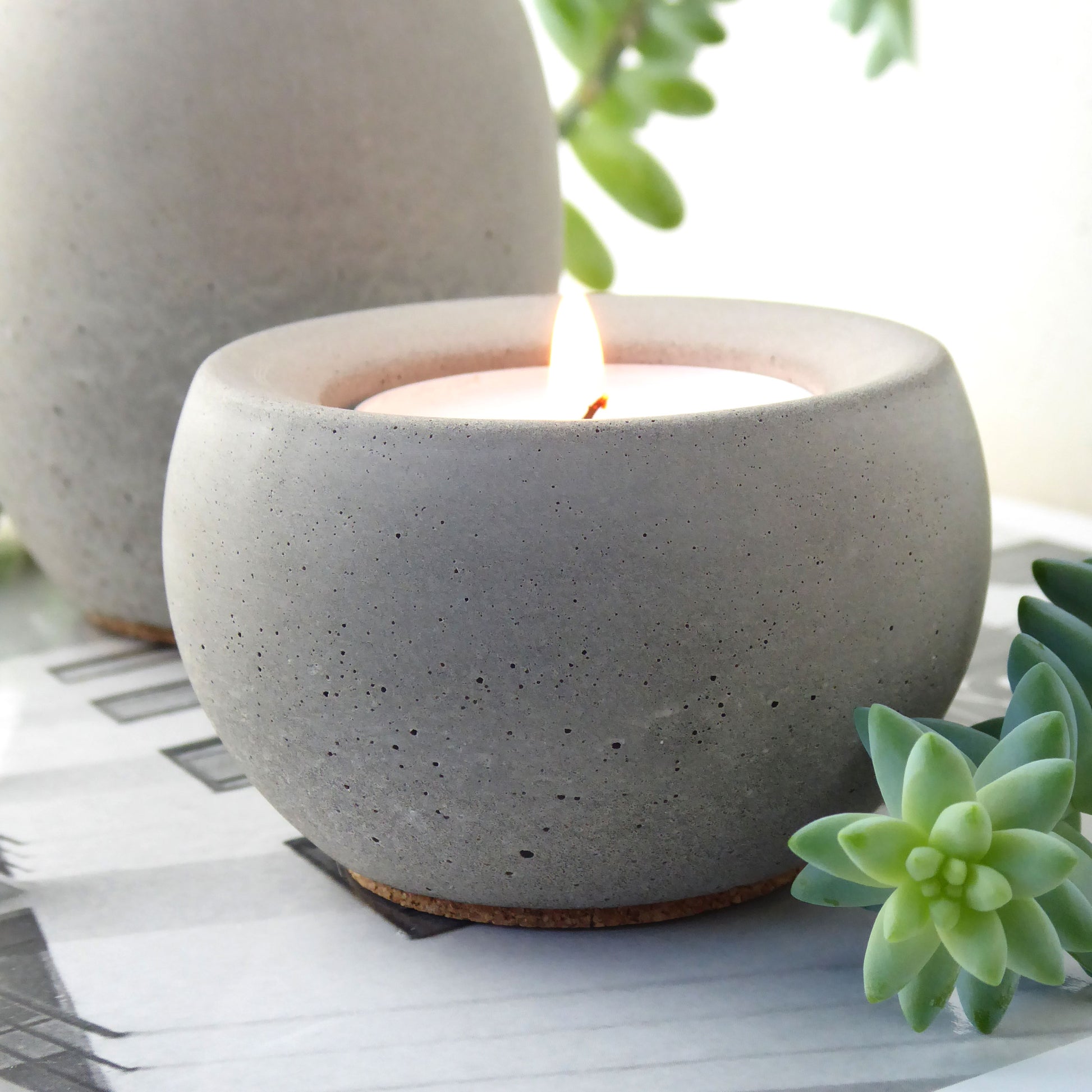 Grey concrete candle holder with a lit candle on a white surface with green plants.