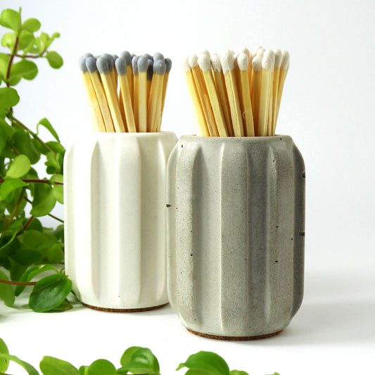 Two concrete match holders in white and grey containing long matches on a white background with greenery.