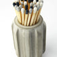 Long matches with black, white, and grey heads in a concrete pot holder with a ridged design on a white background