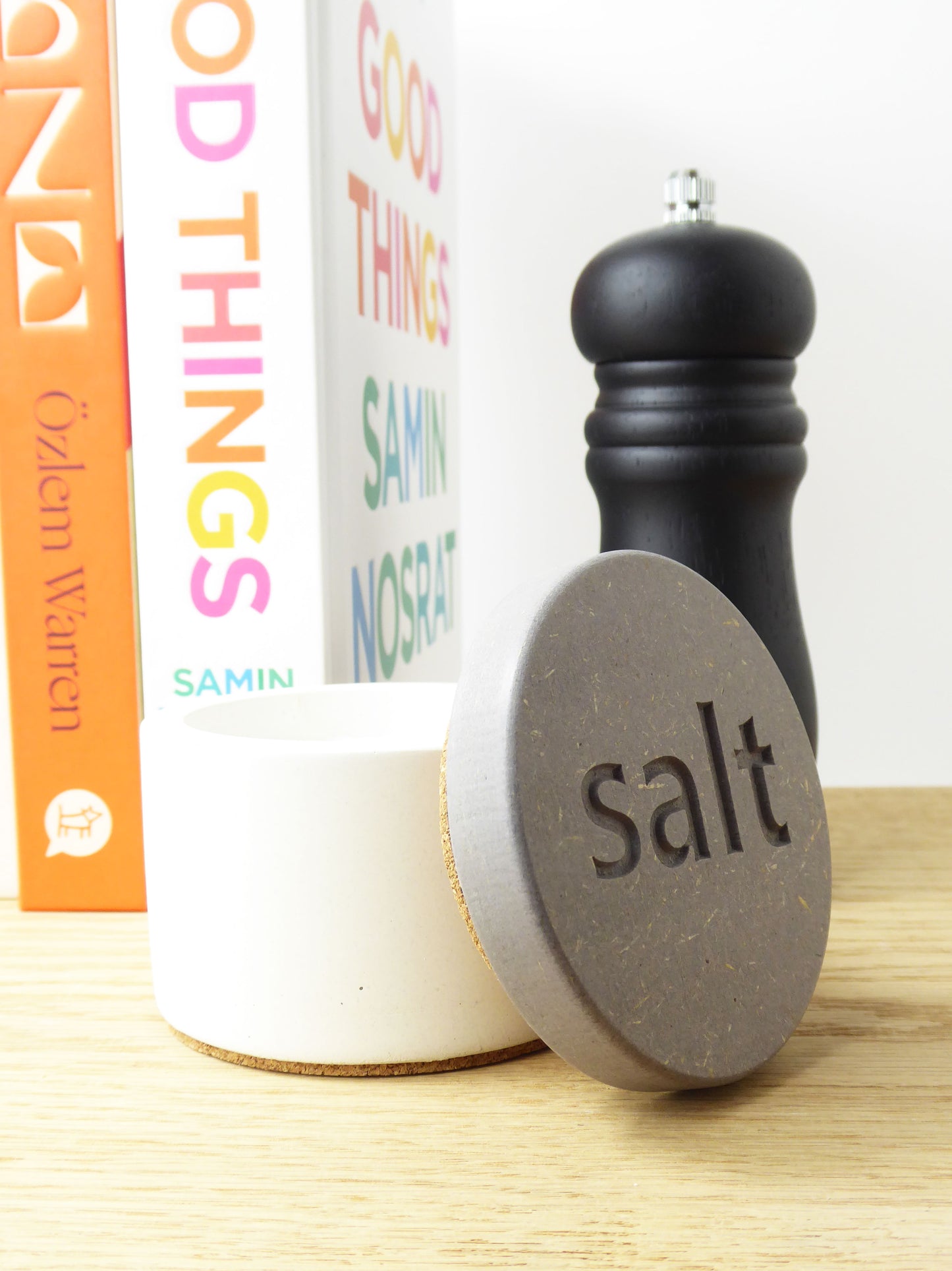 White concrete salt pot for storing salt flakes next to a pepper mill on a kitchen worktop next to some cookbooks