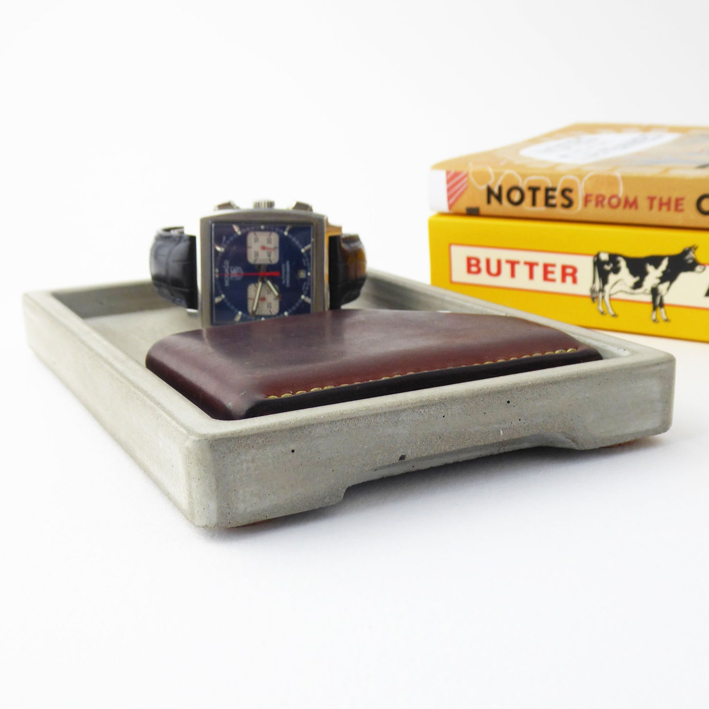 Grey concrete tray with a watch and wallet on a white background next to some books