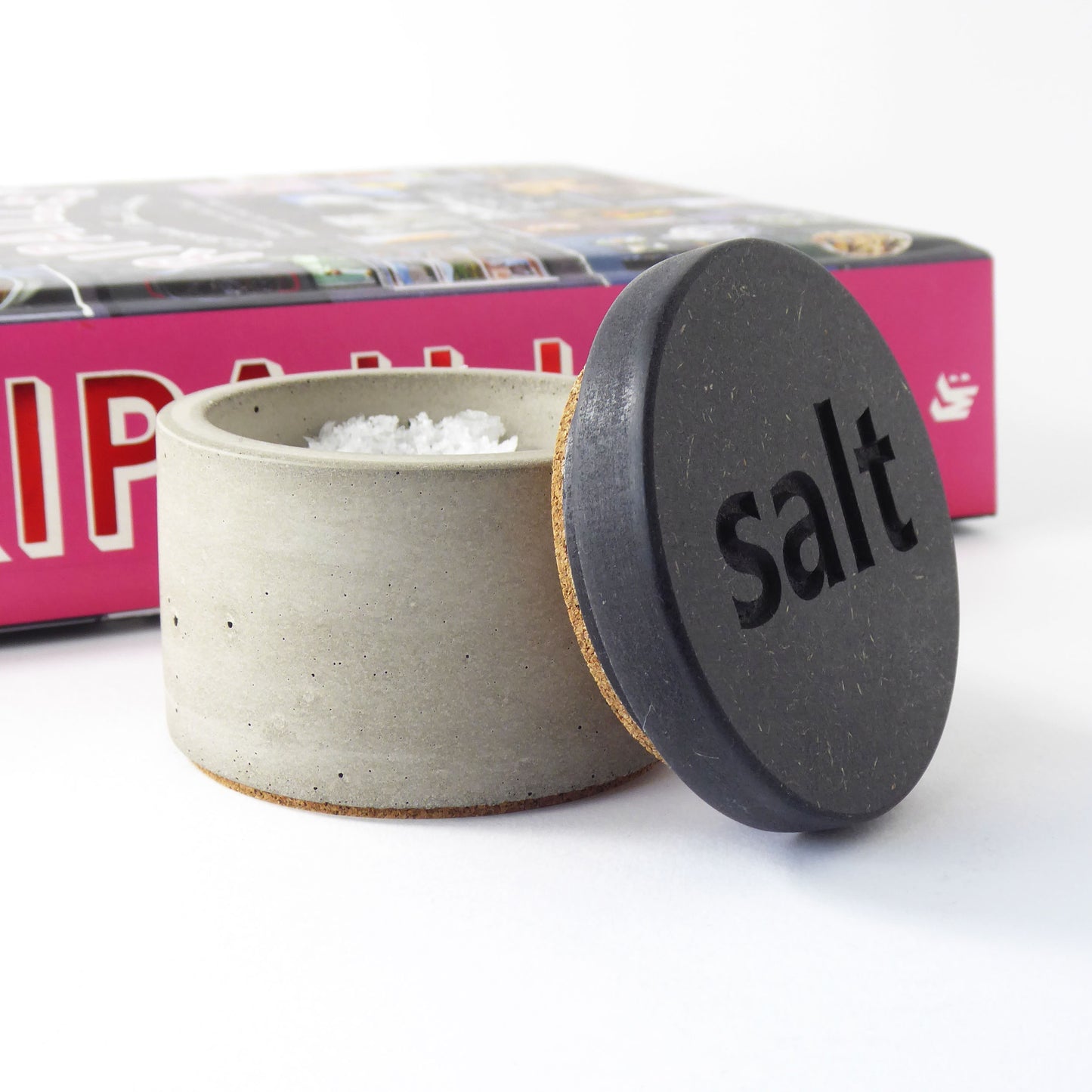 Concrete salt cellar with a black wooden lid engraved with 'salt' on a white background next to a cook book