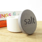 White round concrete salt pot with a grey wooden lid engraved with the word 'salt' on a kitchen worktop next to some cookbooks 