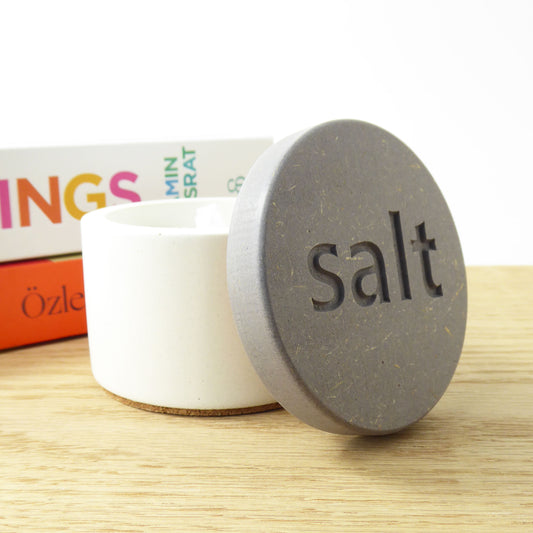 White round concrete salt pot with a grey wooden lid engraved with the word 'salt' on a kitchen worktop next to some cookbooks 