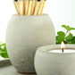 Natural concrete pot with coloured long matches and a matching concrete candle holder with a lit tea light, surrounded by greenery.