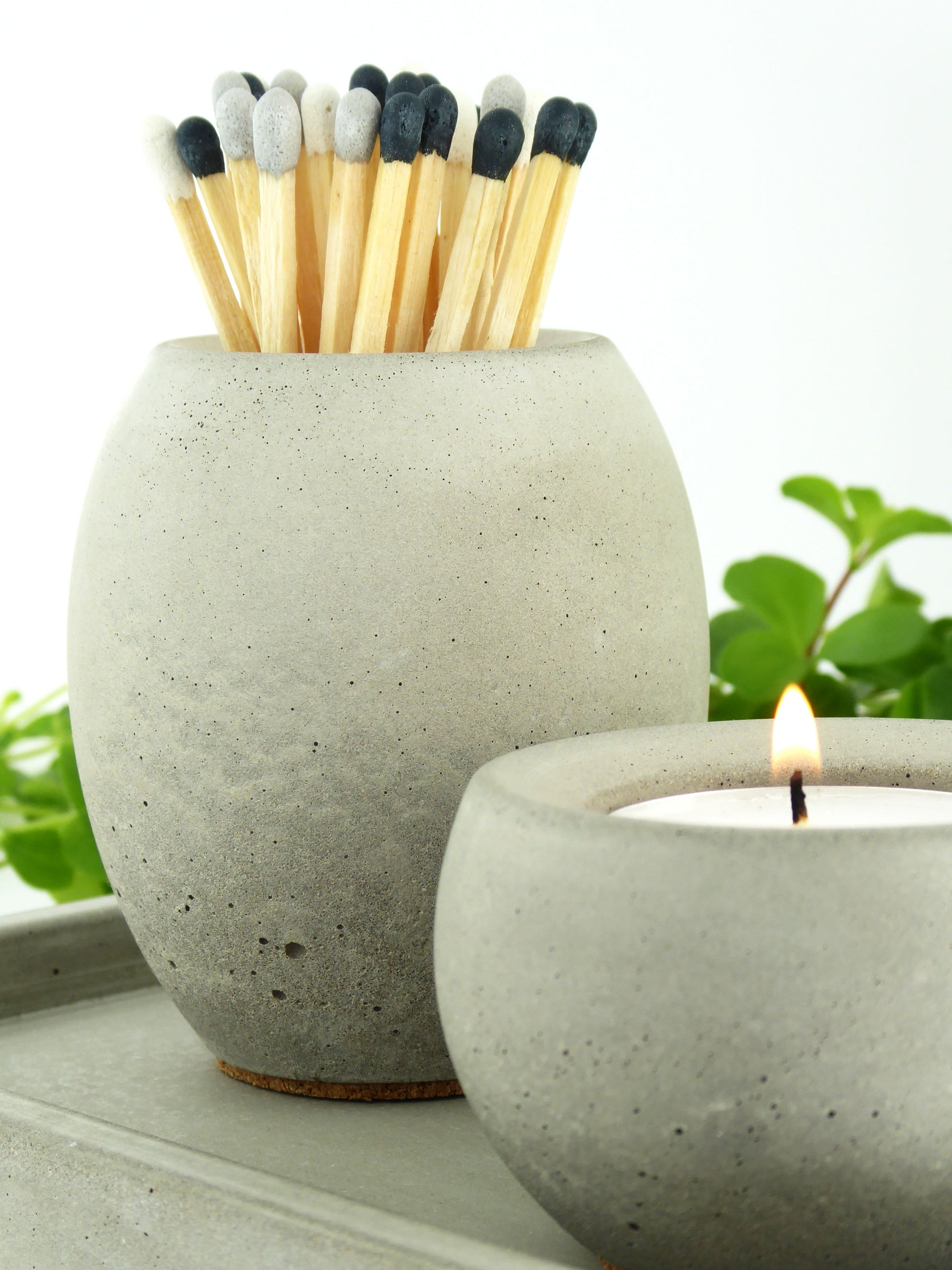 Natural concrete pot with coloured long matches and a matching concrete candle holder with a lit tea light, surrounded by greenery.