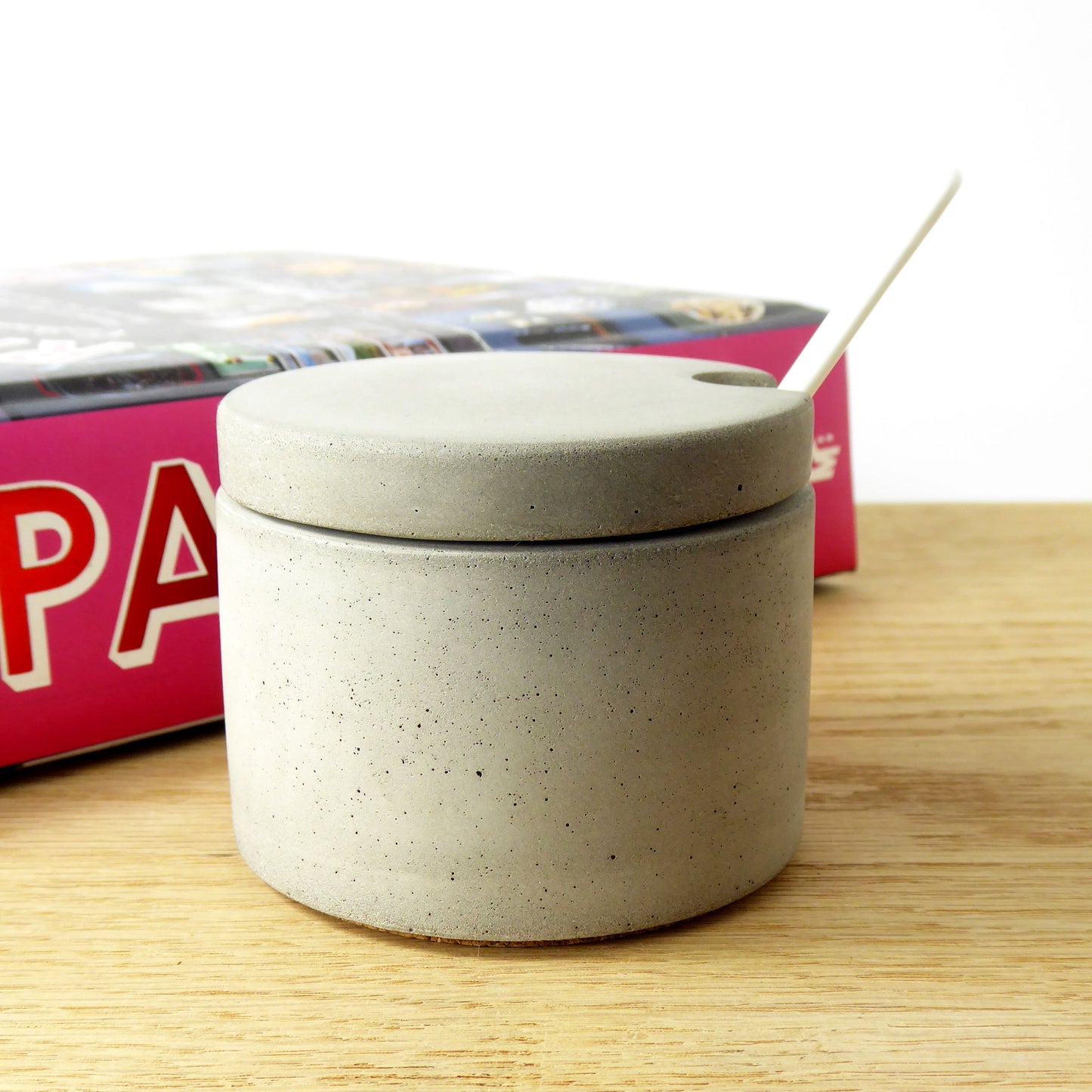 Grey concrete pot with a lid and white spoon on a shelf next to cookbooks
