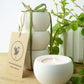 Gift set of 3 white ceramic candle holders with a lit candle, surrounded by greenery on a wooden surface.