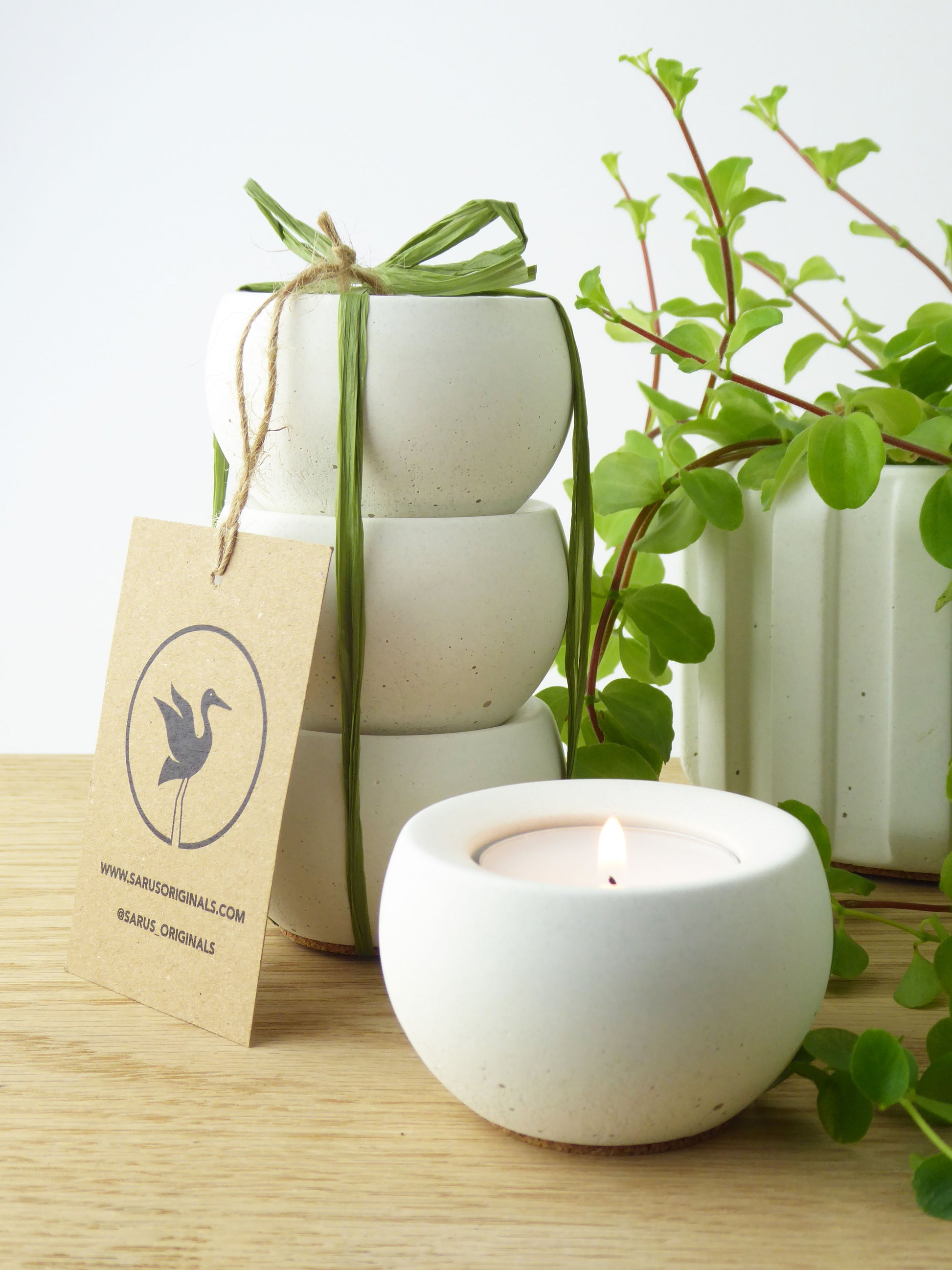 Gift set of 3 white ceramic candle holders with a lit candle, surrounded by greenery on a wooden surface.