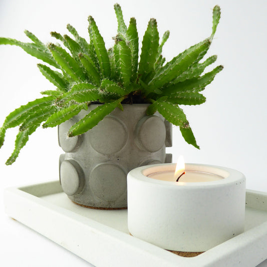 Concrete planter with a green succulent plant and a white candle holder with a lit candle on a white rectangular tray.
