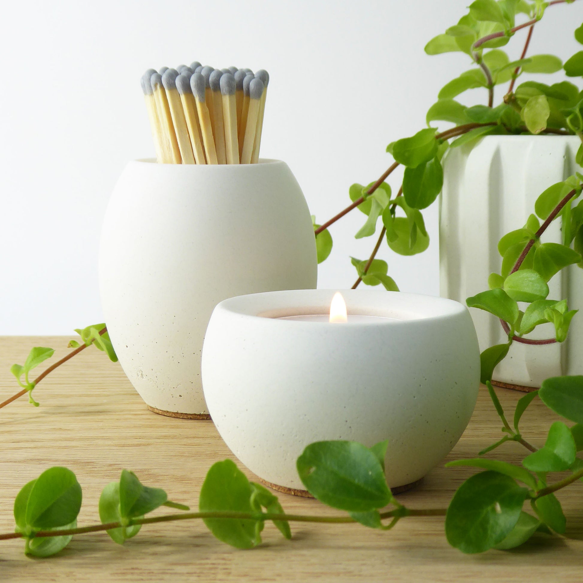 White concrete candle holder with a lit candle and a matching concrete pot holding long matches, surrounded by green leaves on a light wooden surface.