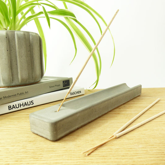 long grey concrete incense holder on a wooden shelf next to books and a houseplant