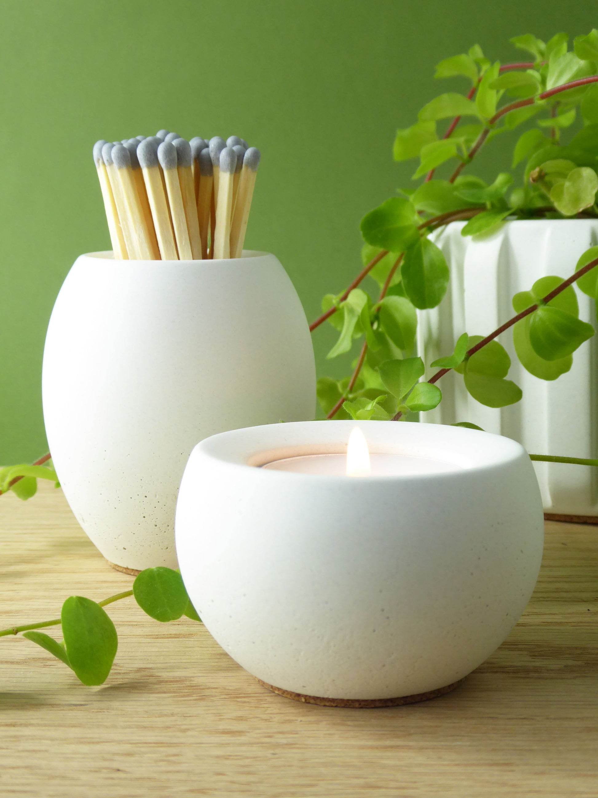 White concrete match pot holder filled with long matches and a matching concrete candle with a lit tea light inside on a wooden shelf with greenery.