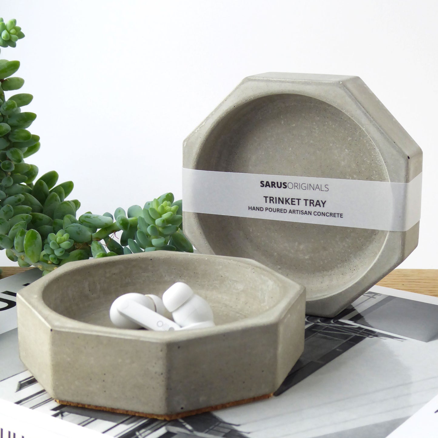 Grey concrete trinket tray containing ear buds on a bedside table next to books and a houseplant