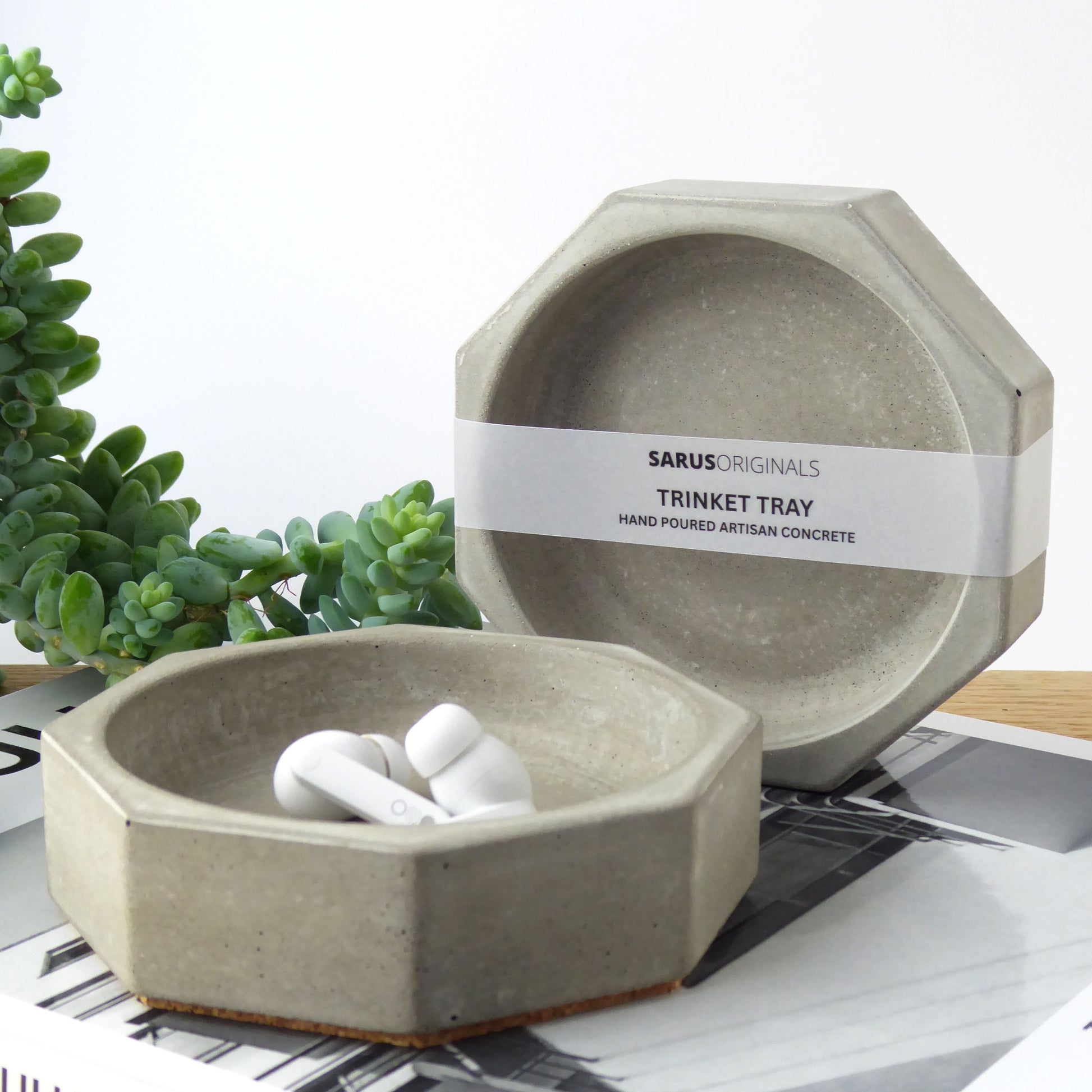 Grey concrete trinket tray containing ear buds on a bedside table next to books and a houseplant