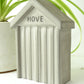 Raw grey concrete beach hut-shaped object with 'HOVE' engraved in the design on the front styled next to a houseplant on a white surface.