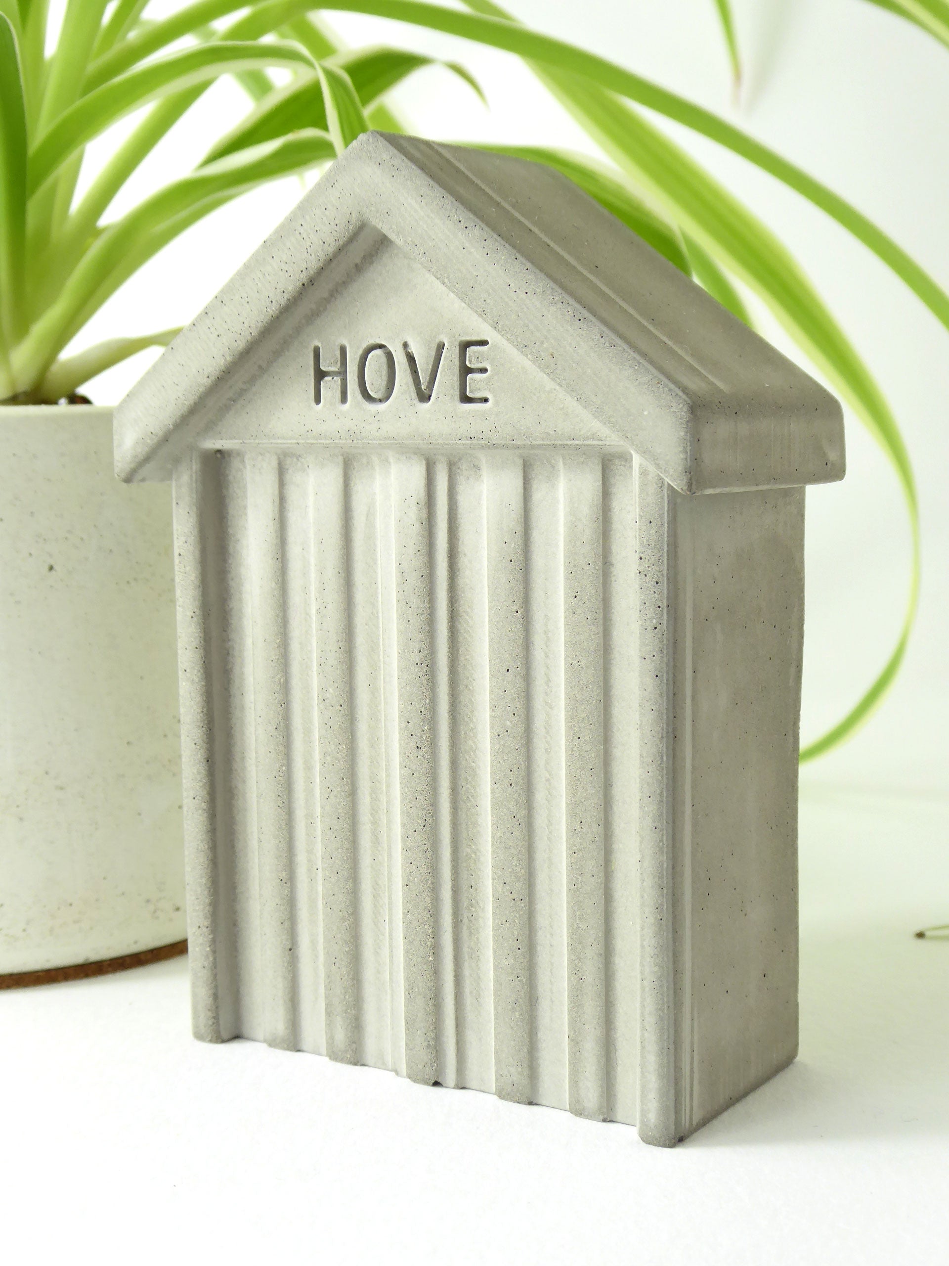 Raw grey concrete beach hut-shaped object with 'HOVE' engraved in the design on the front styled next to a houseplant on a white surface.
