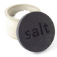 Grey concrete pot containing salt flakes with a black wooden lid with the word 'salt' engraved on it on a white background