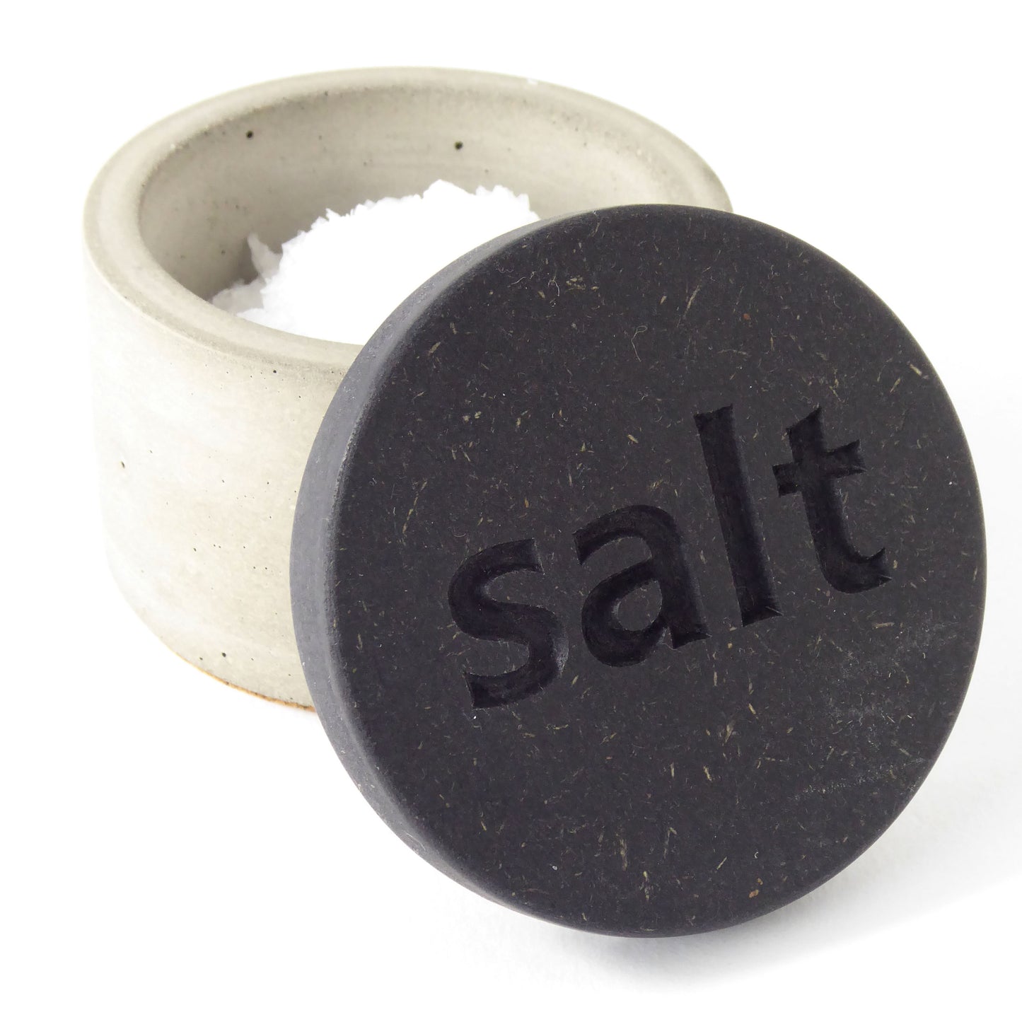 Grey concrete pot containing salt flakes with a black wooden lid with the word 'salt' engraved on it on a white background