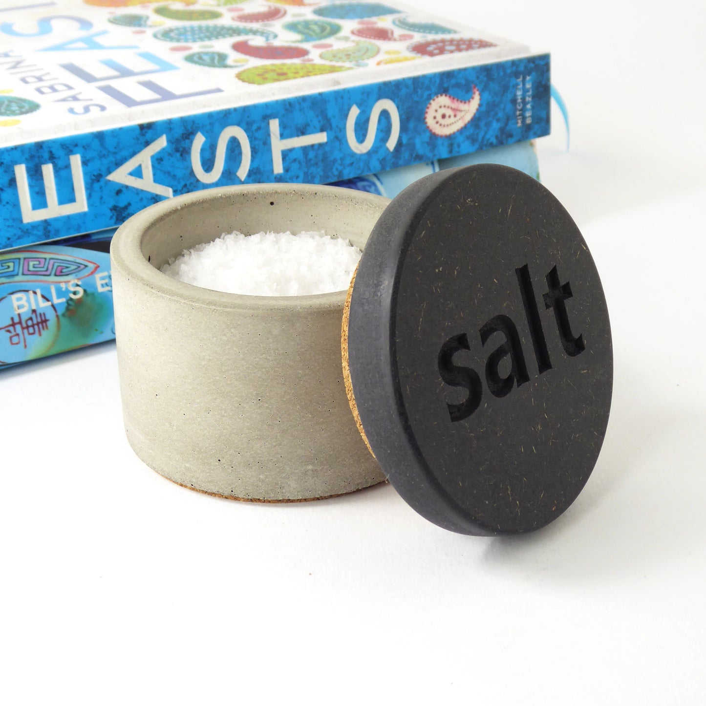 Salt container made of grey concrete with a black wooden lid engraved with the word 'salt' on a white background next to cookbooks