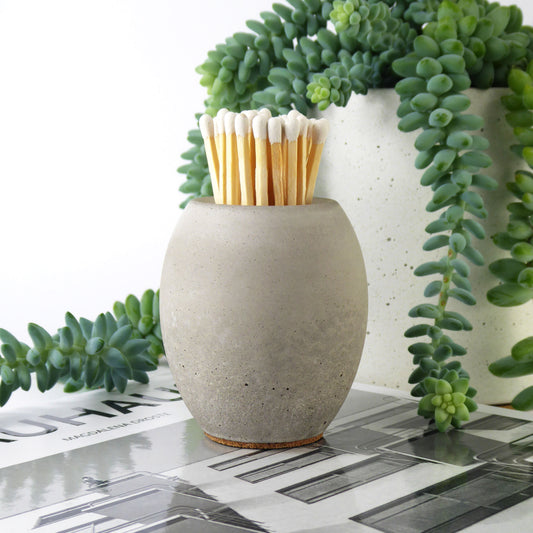 Grey concrete match holder with long matches with coloured tips on a surface with greenery in the background