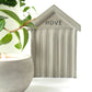 Small concrete beach hut-shaped ornament with 'HOVE' embossed in the concrete styled next to a concrete candle and a houseplant