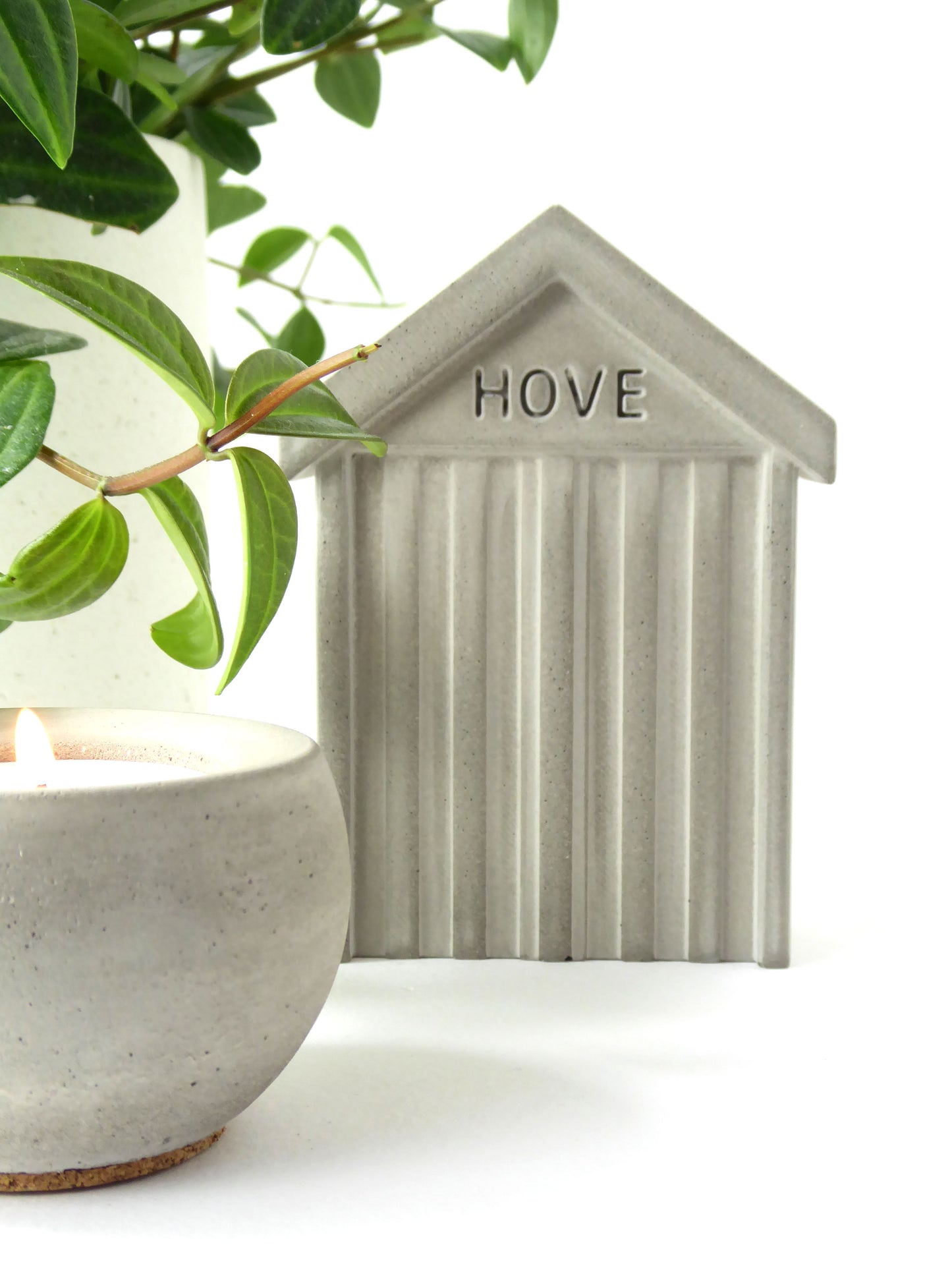 Small concrete beach hut-shaped ornament with 'HOVE' embossed in the concrete styled next to a concrete candle and a houseplant