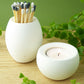 White concrete match pot with coloured matches and a small white concrete candle holder on a wooden shelf with greenery.