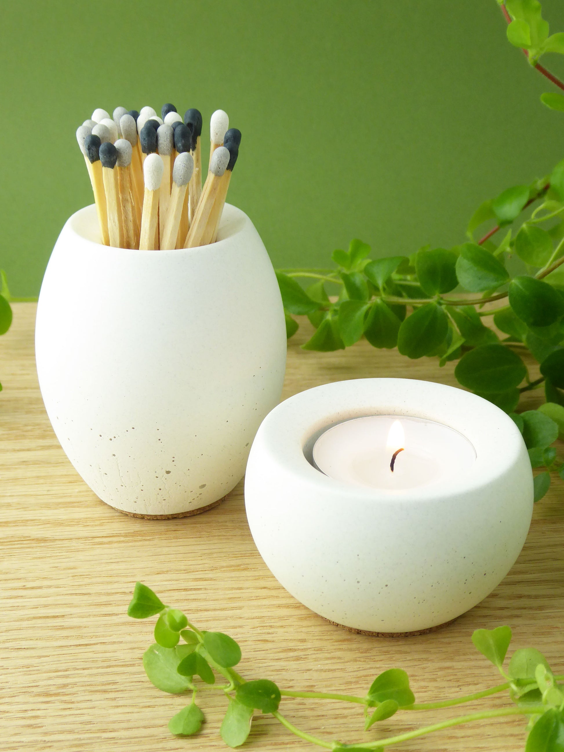 White concrete match pot with coloured matches and a small white concrete candle holder on a wooden shelf with greenery.