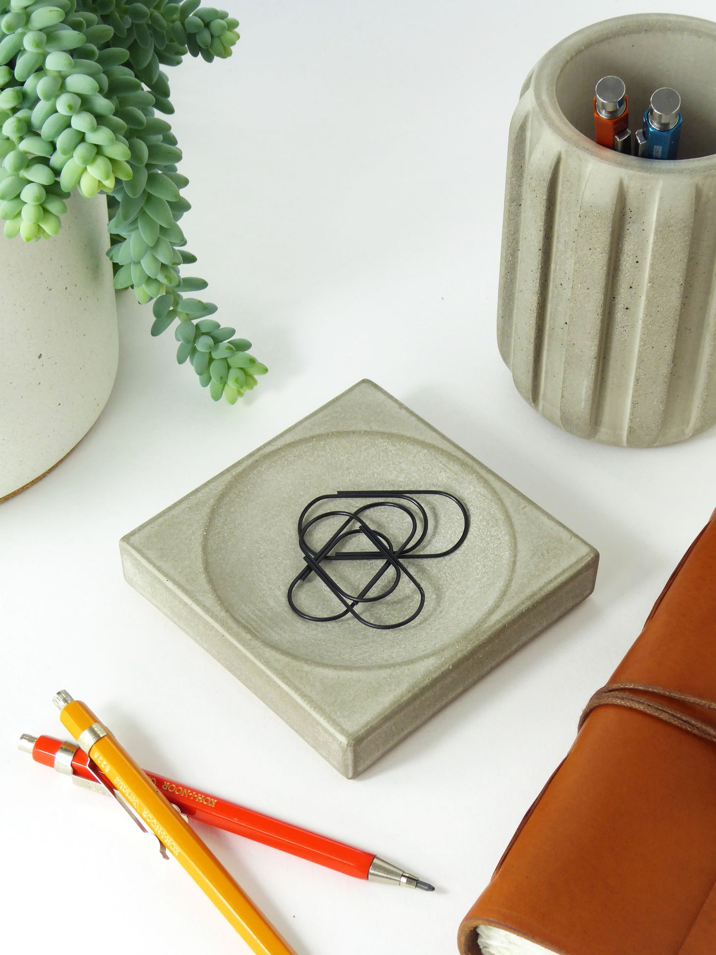 Concrete desk organiser with paperclips in it on a desk next to, pencils in a matching concrete pen pot, and a notebook on a white surface.