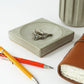Grey concrete decorative tray with metal paperclips, orange and red pens, and a leather-bound notebook on a desk