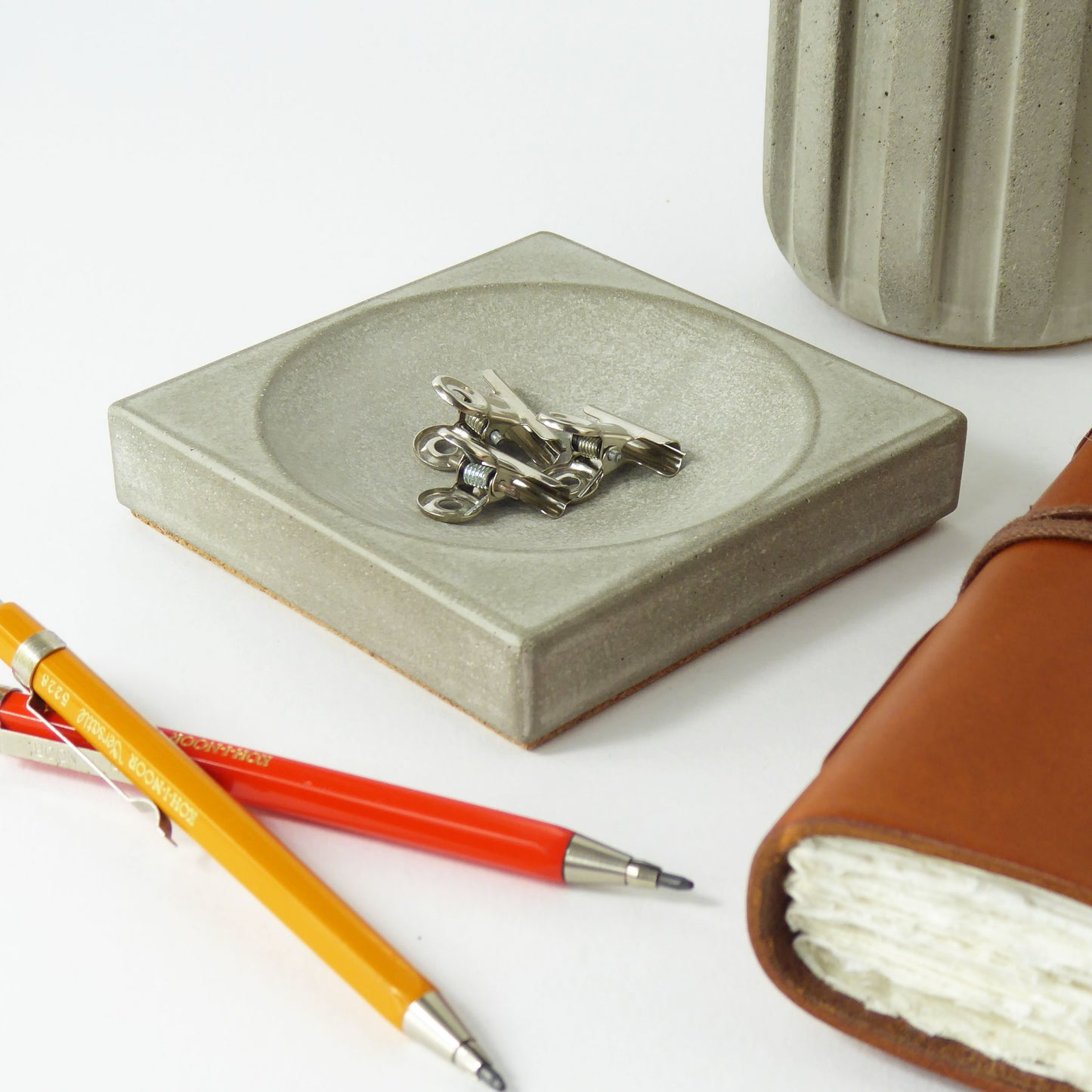 Grey concrete decorative tray with metal paperclips, orange and red pens, and a leather-bound notebook on a desk