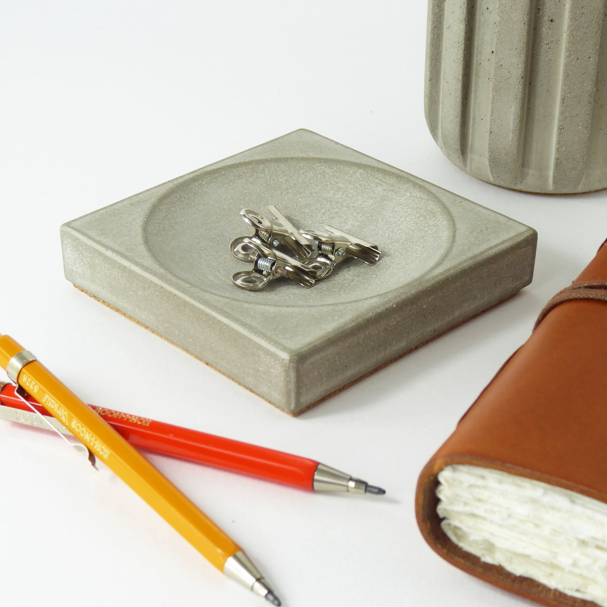 Grey concrete decorative tray with metal paperclips, orange and red pens, and a leather-bound notebook on a desk