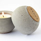 Concrete candle holder with cork base and SARUS logo next to a lit candle on a white background