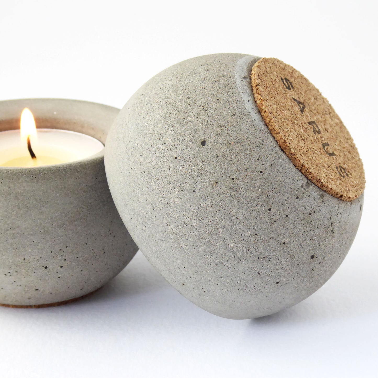 Concrete candle holder with cork base and SARUS logo next to a lit candle on a white background