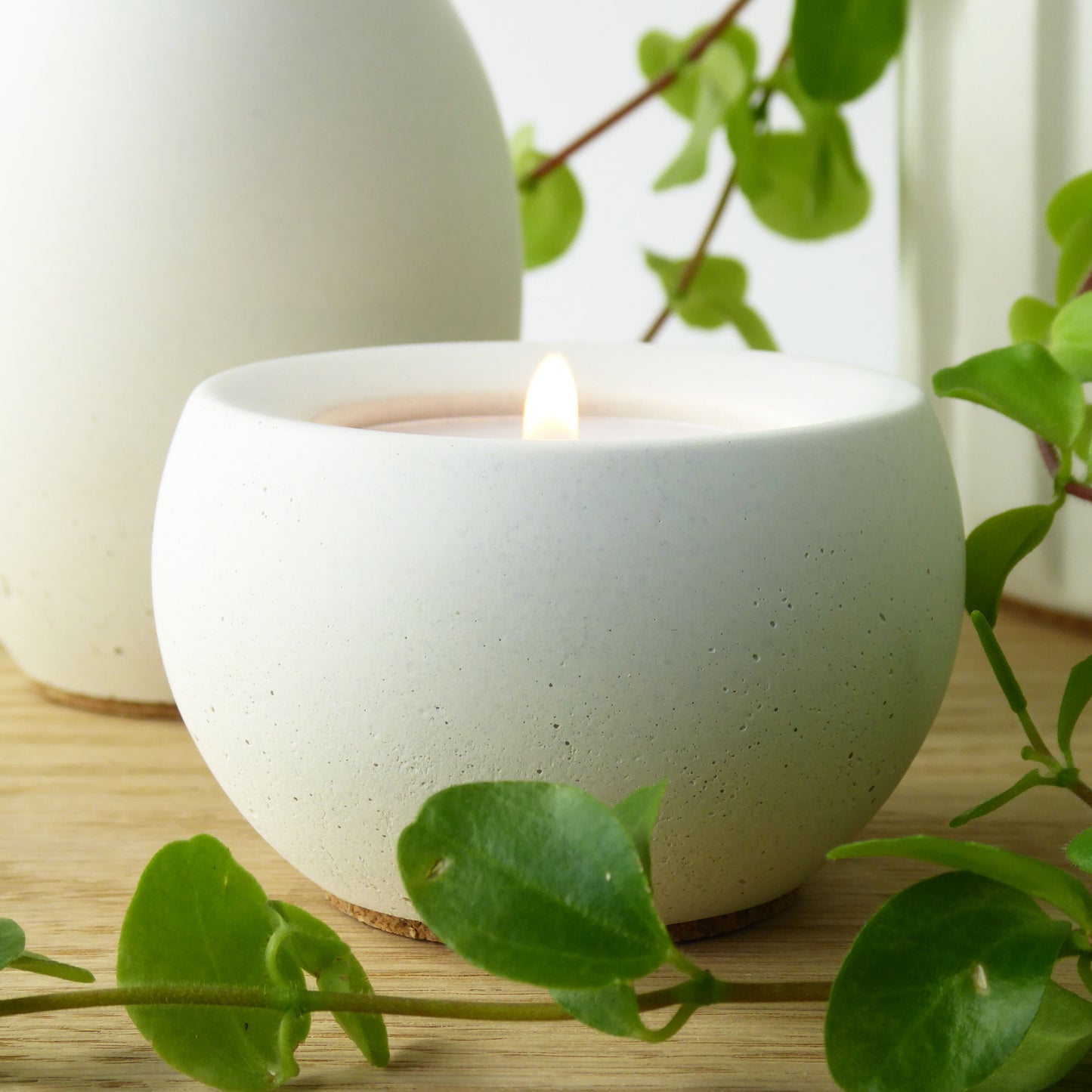 White concrete candle holder with a lit candle on a wooden table with green leaves.
