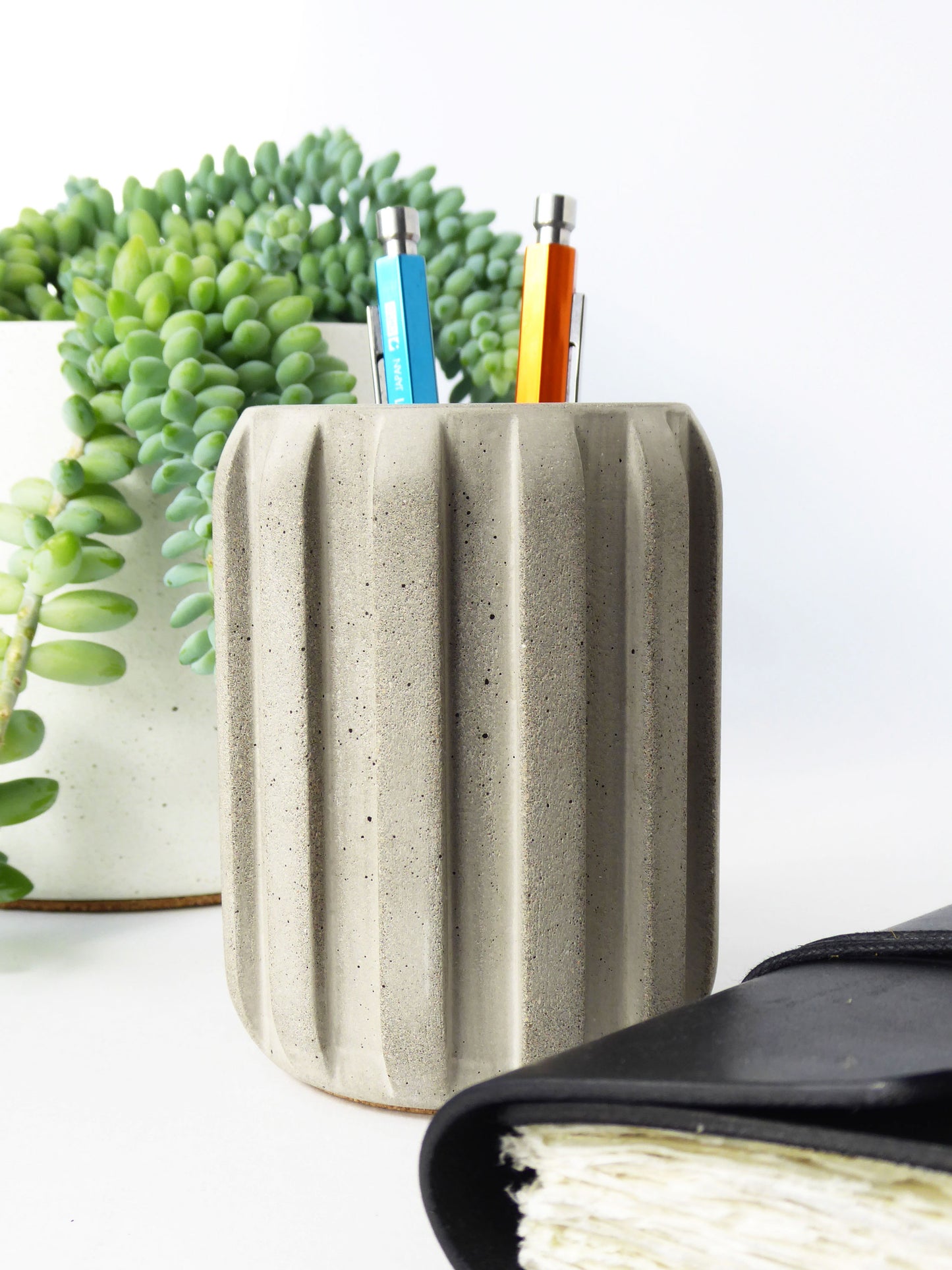 Concrete pencil holder with pens and a plant on a white surface next to a leather journal