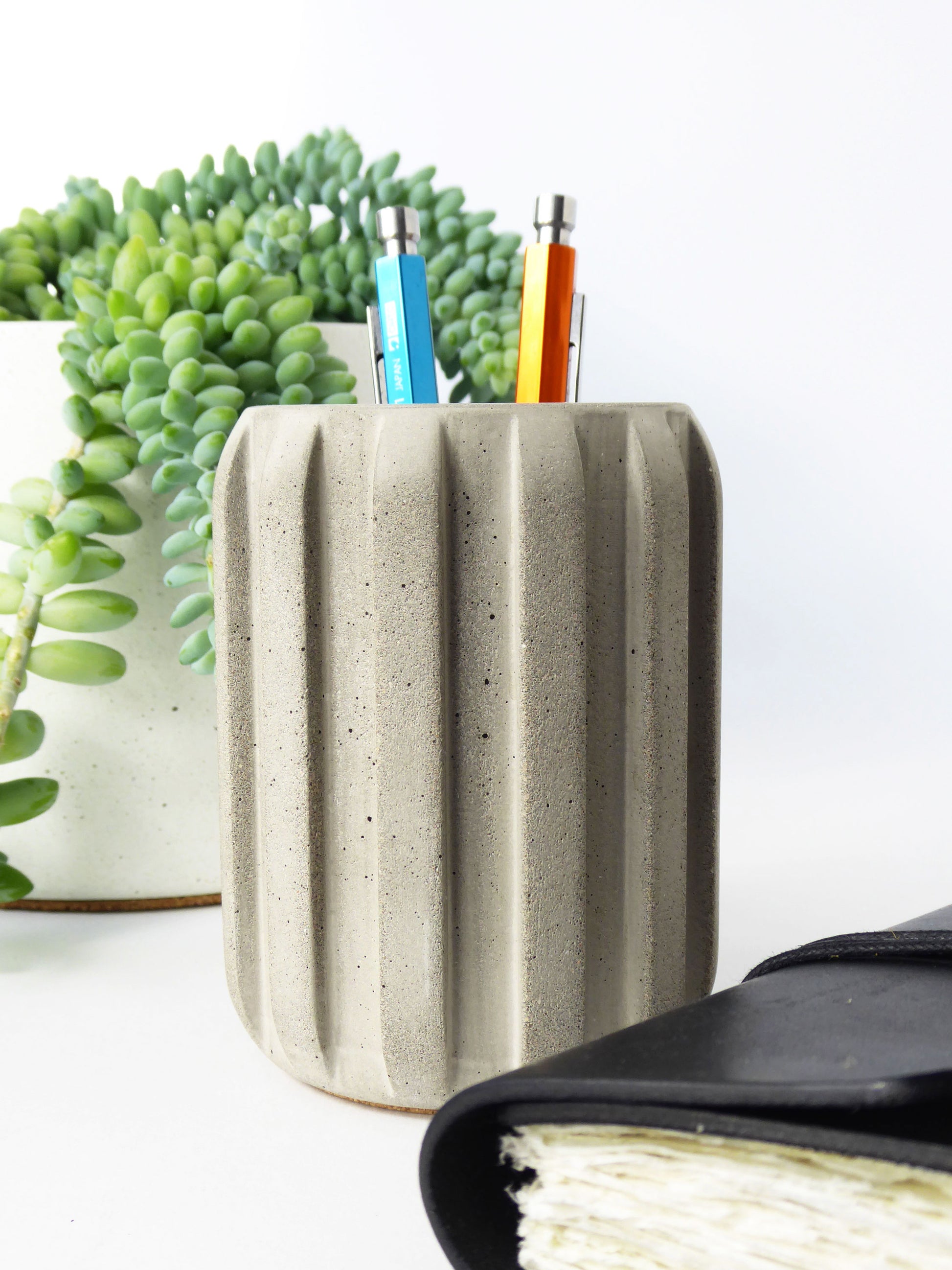 Concrete pencil holder with pens and a plant on a white surface next to a leather journal