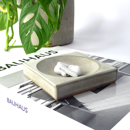 Grey concrete square tray holding white wireless ear pods with a book and plant pot