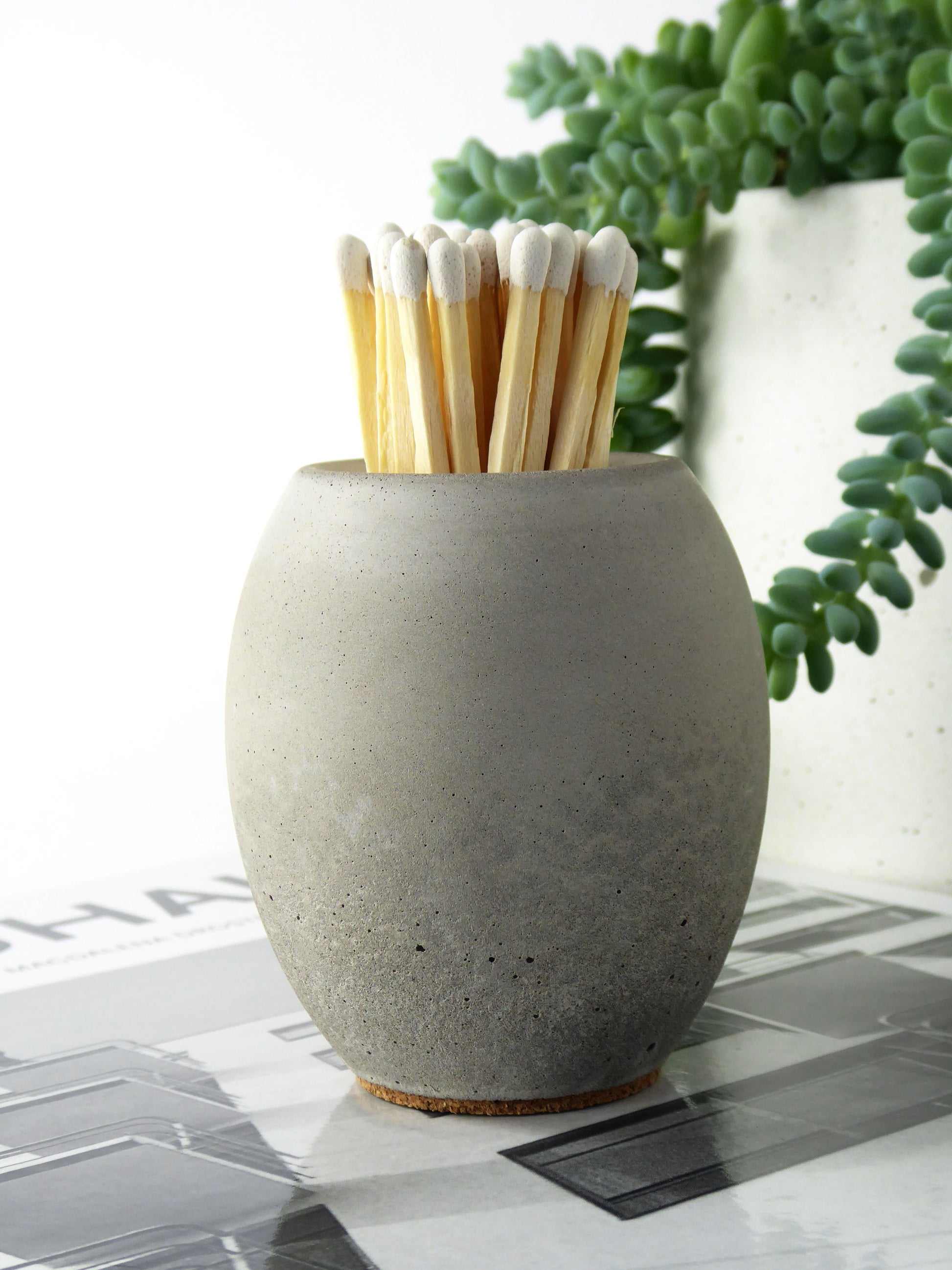 Grey concrete match stick holder with long 10cm matches on a table with plants in the background