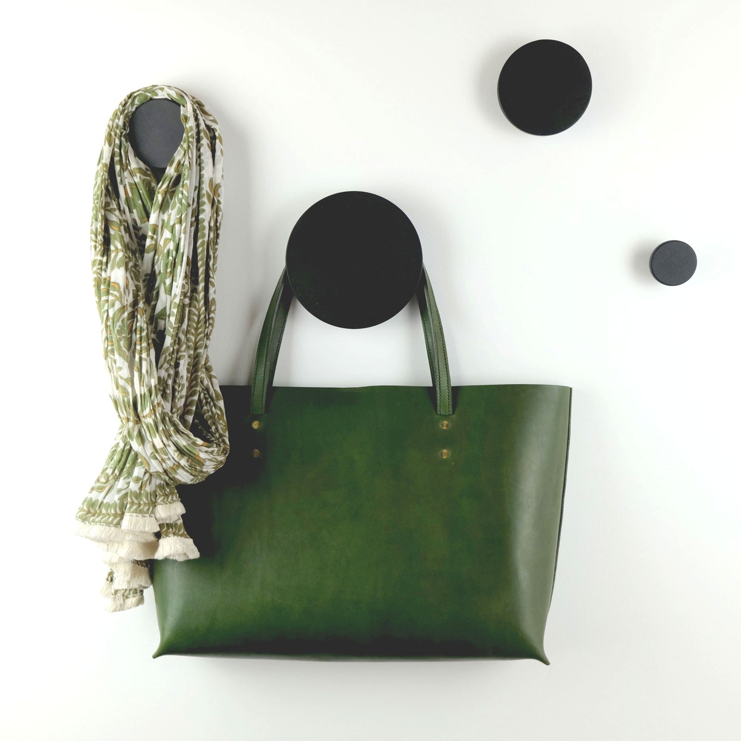 A set of black decorative wooden wall hooks of five different sizes displayed on a white wall, with one hook holding a green leather bag and another holding a multicoloured green scarf.