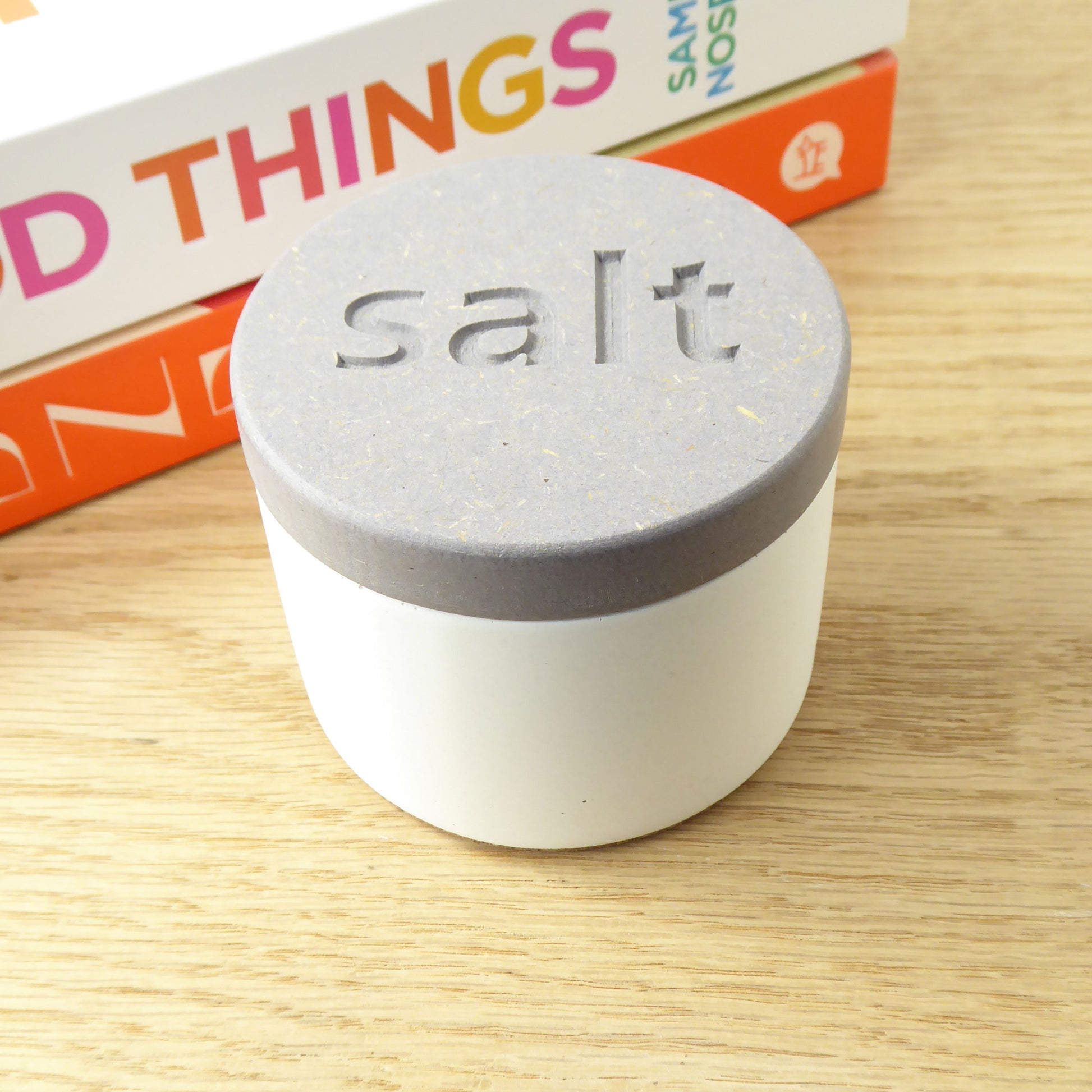 White concrete pot with a gray wooden lid engraved with the word 'salt' on a shelf next to some cookbooks