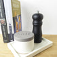 White concrete salt pot next to a black pepper grinder on a white rectangular concrete tray styed on a kitchen shelf next to some cook books