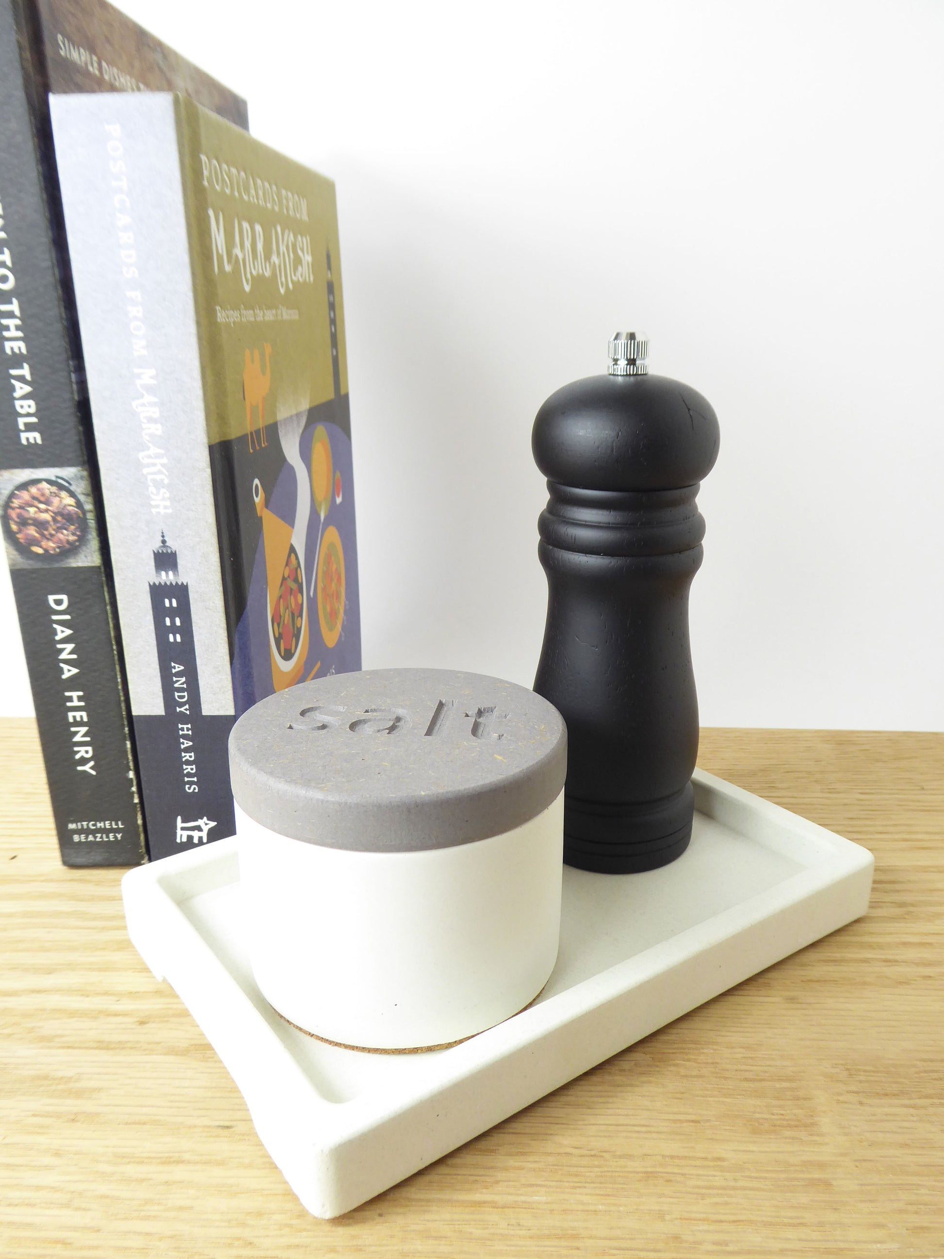 White concrete salt pot next to a black pepper grinder on a white rectangular concrete tray styed on a kitchen shelf next to some cook books