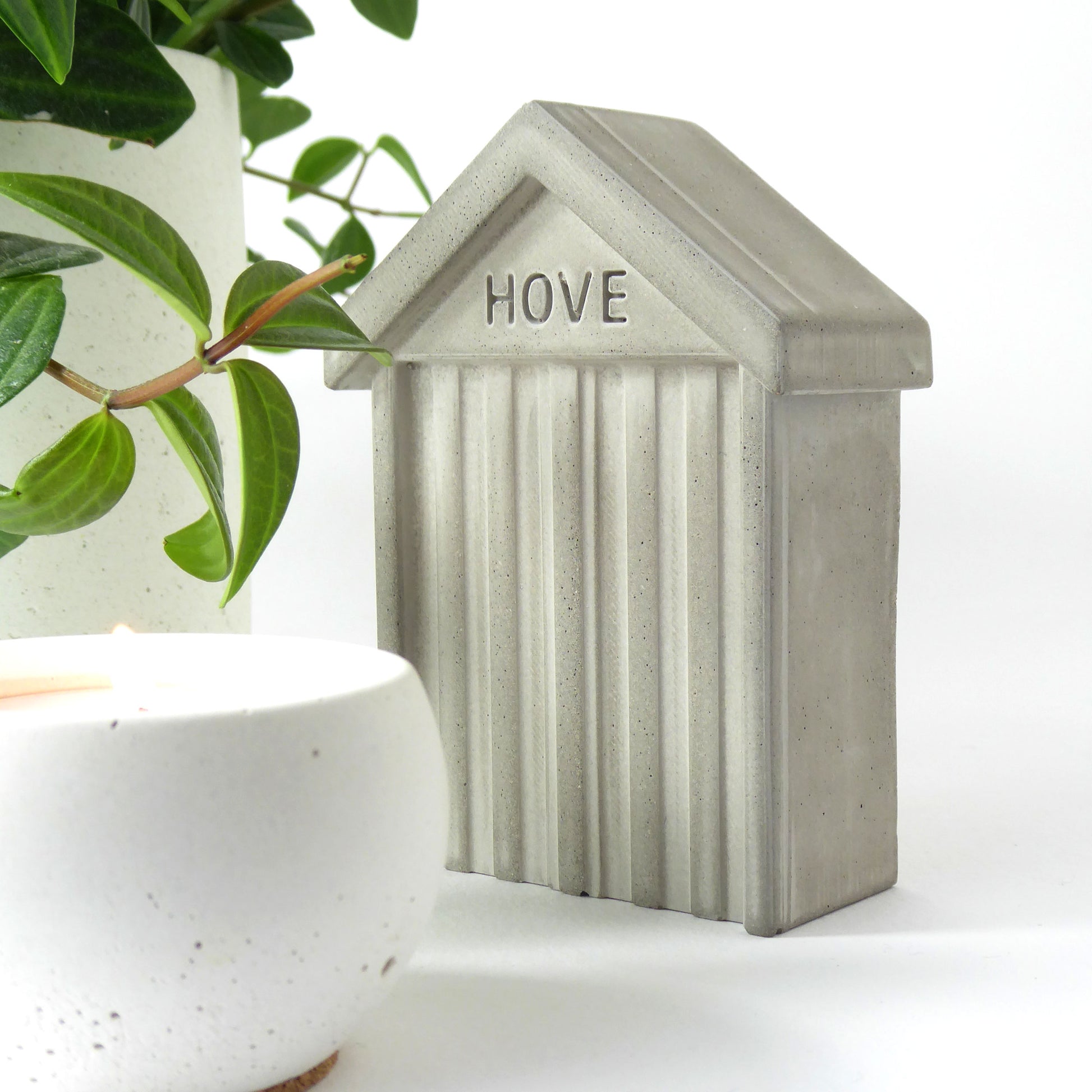 Small concrete beach hut shaped collectible with HOVE embossed in the design on the front next to a white concrete candle and houseplant