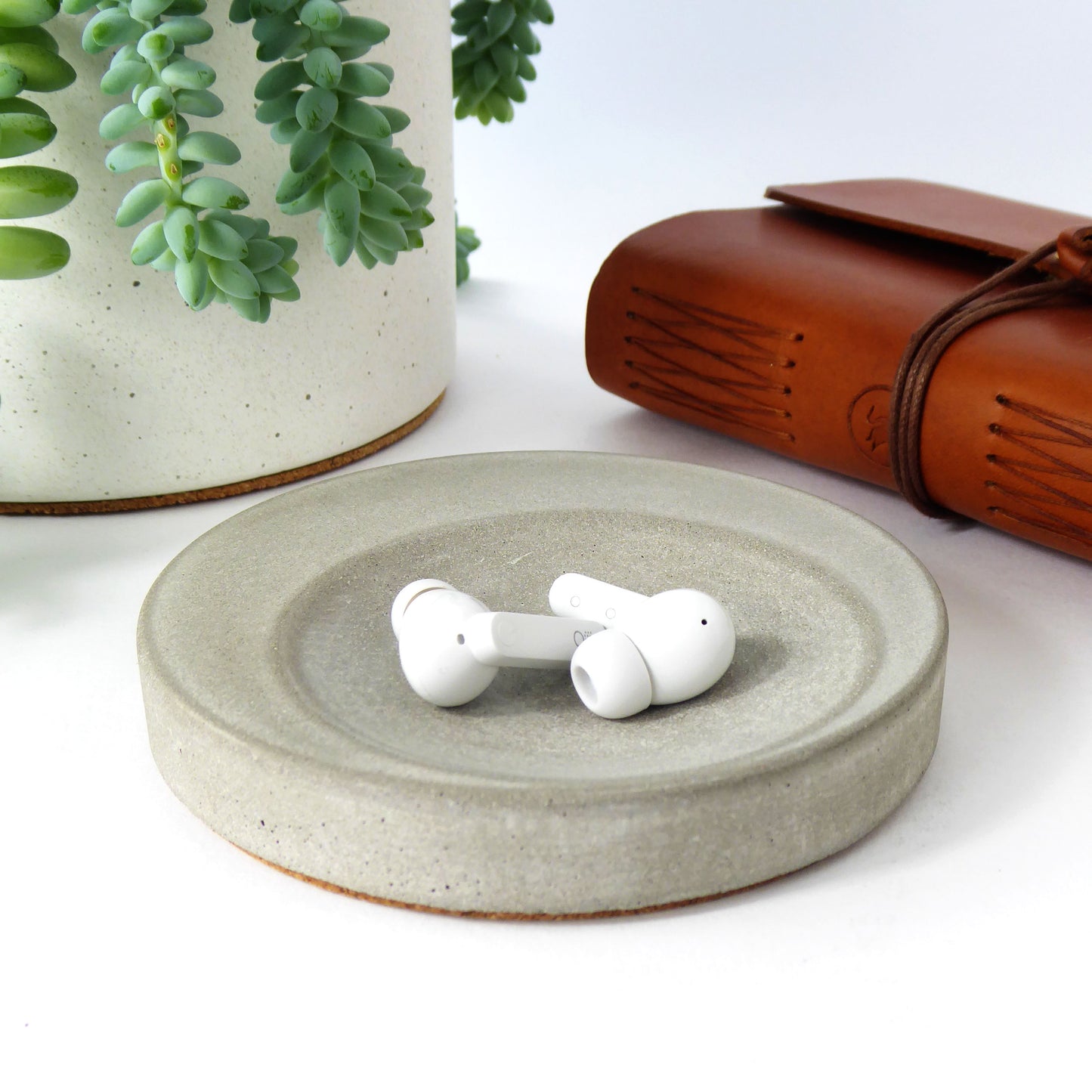 White wireless earbuds in a round grey concrete decorative tray with a plant and brown leather notebook in the background.