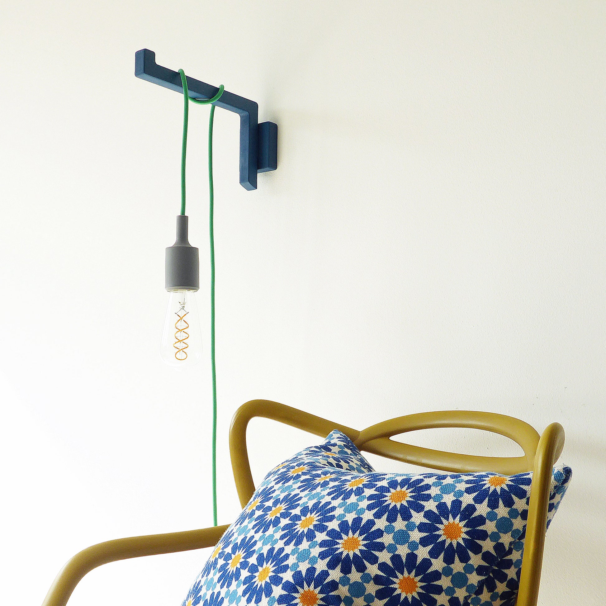 Wall-mounted blue wooden lamp hook with a green plug in pendant cord wrapped around the arm styled above a yellow chair with a blue floral cushion.
